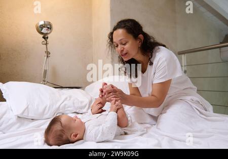 Happy young adult multi ethnic woman, a delightful affectionate loving mother caring of her newborn baby boy, stroking and kissing his little feet and Stock Photo