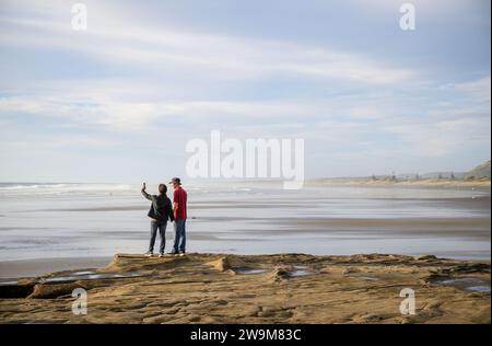 Couple taking photos using smartphone at Muriwai Beach. Auckland. Stock Photo
