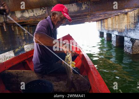 Cabimas, Venezuela. 28th Dec, 2023. A fisherman holds a fish while fishing below an old oil pump. After being exploited for intense oil extraction for decades, Lake Maracaibo has become heavily polluted due to the neglect and lack of maintainance of the old pipes and infrastructure. This has caused a severe decline in fish stock, with the appearance of bacterias and toxic algae on the water surface, damaging the lives of the people living on its shores who rely on fishing for a living. Credit: SOPA Images Limited/Alamy Live News Stock Photo