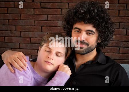 This image portrays a moment of tenderness, with a woman resting her head on a man's shoulder, eyes closed in comfort or perhaps sleep. The man, with curly hair and a black shirt, looks into the camera with a contented smile, suggesting a sense of protection and affection. They are seated against a backdrop of a rustic brick wall, adding texture and a sense of solidity to the scene. Comfort in Embrace. High quality photo Stock Photo