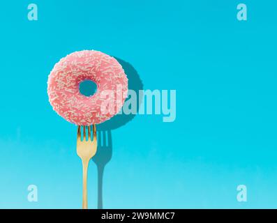Yummy pink glazed donut pricked on a fork on bright light blue background. Creative art. Contemporary style. Minimal concept of food with copy space. Stock Photo