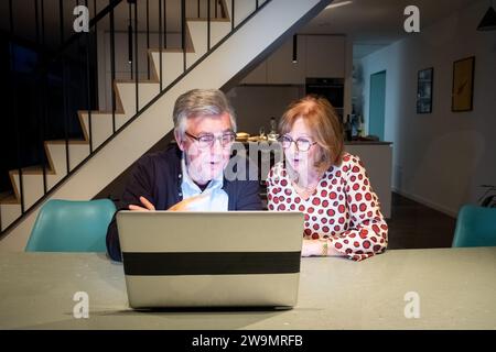 This image captures an elderly couple as they concentrate on a laptop screen. The man, clad in a dark jacket and glasses, shows a look of focus and perhaps discovery. Beside him, the woman in a polka-dot top appears equally absorbed, her posture indicating active participation. They are situated in a contemporary kitchen that opens into a dining area, with the sleek lines of a staircase in the background, exemplifying a modern, connected lifestyle. Senior couple Navigating the Internet Together. High quality photo Stock Photo