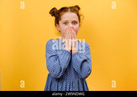 Omg. Portrait of surprised preteen girl child opening mouth and touching face, looking at camera in shock, posing isolated over plain yellow color bac Stock Photo