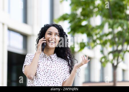 Young beautiful woman talking on the phone close-up while walking in the city, business woman smiling contentedly, successful Latin American woman with curly hair on a sunny day. Stock Photo