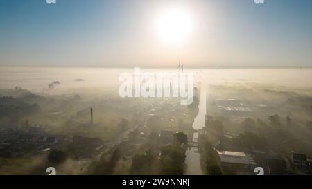 This captivating aerial image showcases a town swathed in a thick blanket of morning mist. The early sun, high in the sky, casts a warm light that gently graces the tops of the mist, creating a surreal landscape. The outlines of buildings and wind turbines emerge from the fog, hinting at human presence in this natural spectacle. The contrast between the clarity above and the foggy veil below invites contemplation of the interaction between civilization and nature. Aerial View of a Town Enveloped in Morning Mist. High quality photo Stock Photo