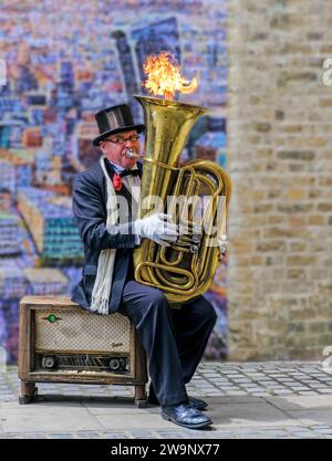 Busker Christopher Werkowicz seated on vintage radio in front of painting London Cityscape by Jimmy C, plays a tuba which shoots out fire. Stock Photo