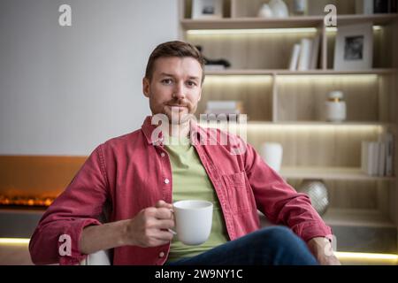 Glad friendly smiling man sitting in cozy flat with cup of tea, drinking, resting, looking at camera Stock Photo