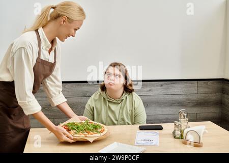 joyful waitress proposing delicious pizza to female client with down syndrome sitting in cozy cafe Stock Photo