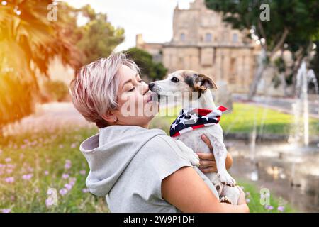 Woman with short grey hair holding and kissed on cheek by small senior dog in park outdoor. Dog is wearing bandana with an American flag. Joy and comp Stock Photo