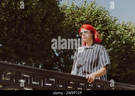 A teenage girl with red hair and sunglasses walks in a city park on a clear summer day, Generation Z Stock Photo