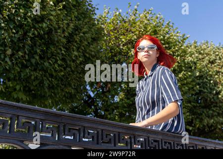 A fashionable teenage girl with red hair and trendy sunglasses enjoys a leisurely walk in the city park on a bright and sunny summer day, showcasing t Stock Photo