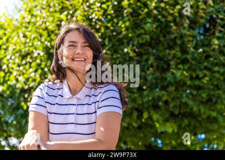 portrait of an attractive smiling middle-aged woman with long hair on a background of greenery in a city park Stock Photo