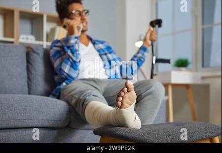 Man talking on phone while sitting on couch, with his injured foot resting on stool Stock Photo