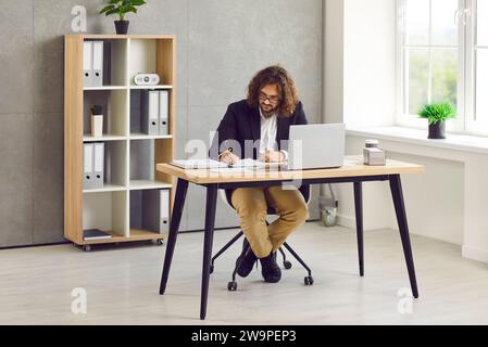 Young stylish business man with long hair working with laptop and writing at the desk in office. Stock Photo