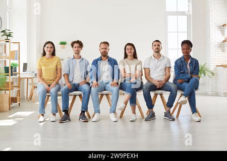 Different multiracial young people in casual clothes sit in row on chairs and look at camera. Stock Photo