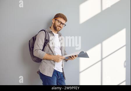 Portrait of happy male student with textbook and backpack standing by grey wall background Stock Photo