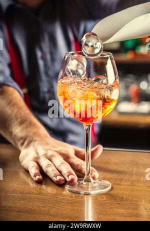 The bartender at the bar prepares the summer cocktail Spritz Veneziano, adds ice cubes to the drink. Stock Photo