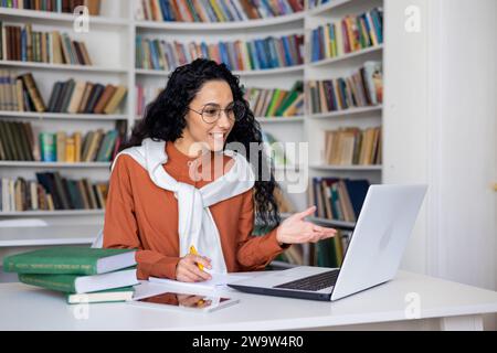 A young Latin American female teacher sits at a desk in a university library and conducts classes remotely, using video communication on a laptop. Stock Photo