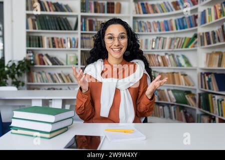 Portrait of a young smiling Latin American female teacher sitting at a desk in a library and teaching online, explaining and gesturing with her hands to the camera. Stock Photo