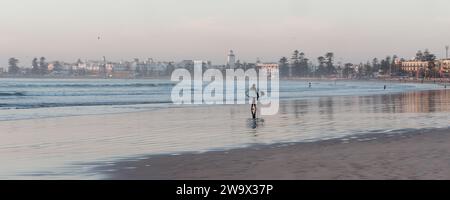 Lone surfer walks out of the water on a sandy beach approaching sunset with the Medina behind in the city of Essaouira, Morocco. December 30, 2023 Stock Photo