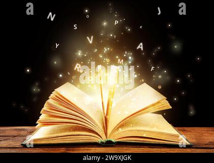 Open book with magic light and glowing letters flying out of it on wooden table against black background Stock Photo