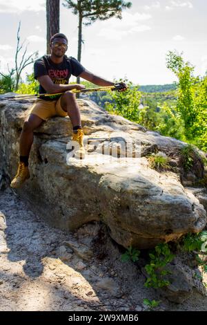A portrait of an African male with hiking gear, sitting on a rock in nature on a hiking trail, holding hiking poles. Stock Photo