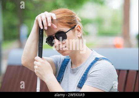 Blind red-haired woman sitting outdoors leaning on a cane.  Stock Photo