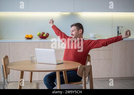 Happy freelancer man stretching arms smiling ending work sitting on kitchen with laptop.  Stock Photo