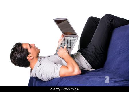 Young attractive man working with computer while laying in awkward position on couch, on white background Stock Photo
