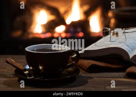 Cup of coffee with open book and blanket with a burning fireplace in background Stock Photo