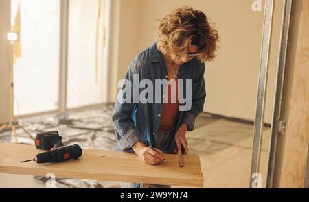 DIY homeowner, a mature woman, measures baseboard in her kitchen for a remodeling project. Using a tape measure, she marks and prepares for the renova Stock Photo