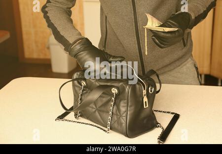 Robber in black outfit and gloves see in opened stolen women bag. The thief takes out the gold and money from a womans handbag in kitchen interior Stock Photo