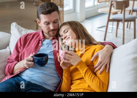 Pleased loving couple enjoying lazy weekend resting at home drinking coffee together on home couch. Stock Photo