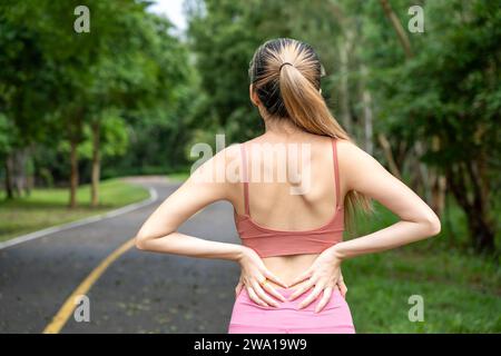 Back view of a long hair woman with back pain putting her hands over her trouble lower back while standing at a running track of a local park Stock Photo