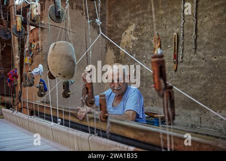 An older man works on horizontal loom in weaving workshop. Kashan, Iran. Stock Photo