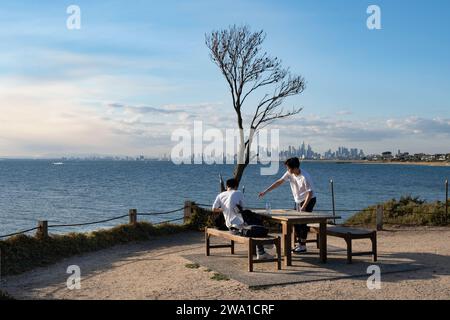 Two Chinese boys drink at a wooden picnic table in Brighton, a suburb of Melbourne on the edge of Hobson Bay with a view of the city skyline Stock Photo
