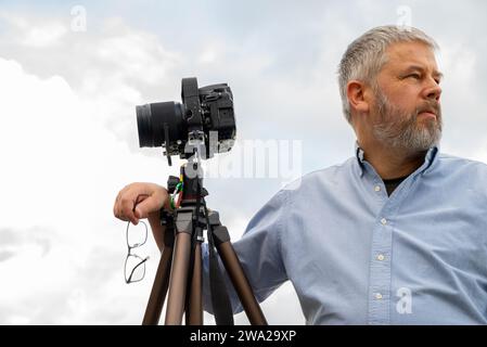 Photographer at work, gray hair and full beard, 56 years old, light shirt, glasses set off in hand, 35mm SLR camera on tripod, gray overcast sky Stock Photo
