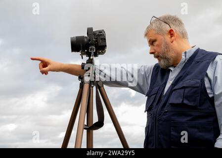 Photographer at work, gray hair and full beard, 56 years old, dark blue photographer's vest, glasses pushed up, pointing in the direction of the camer Stock Photo