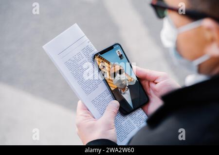 Man on a video call during COVID-19, showcasing pandemic life with a phone in hand, paper, mask, and reflection in glasses Stock Photo