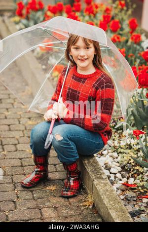 Outdoor spring portrait of adorable kid girl holding transparent umbrella, wearing red plaid pullover and rain boots, child playing outside on a rainy Stock Photo