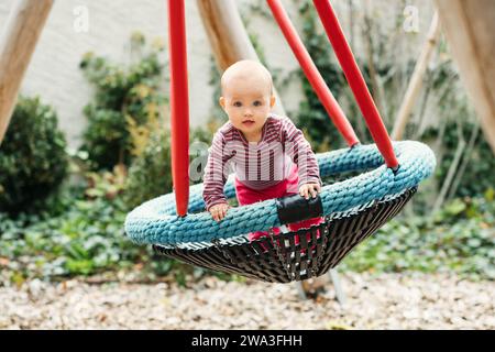 Baby girl having fun in the park, 9-12 months old kid playing in the big swing, summer playground, activities for children Stock Photo