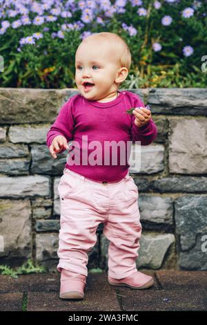 Outdoor portrait of adorable 9-12 month old baby girl playing with purple flowers, wearing pink trousers, leather boots and purple body Stock Photo