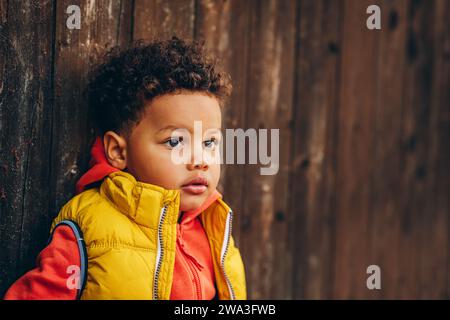 Outdoor portrait of adorable toddler boy posing outside against brown wooden background, wearing orange hoody jacket any bright yellow vest coat Stock Photo