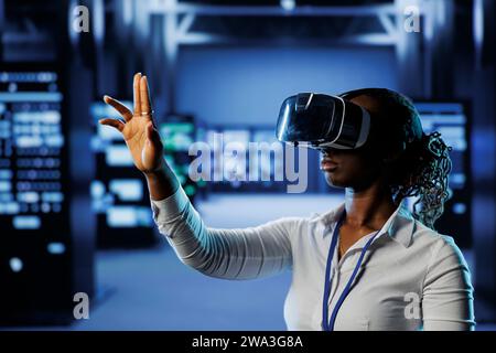 Computer scientist in data center uses virtual reality to prevent system overload. African american VR headset user in server room ensuring enough network bandwidth for smooth operations Stock Photo