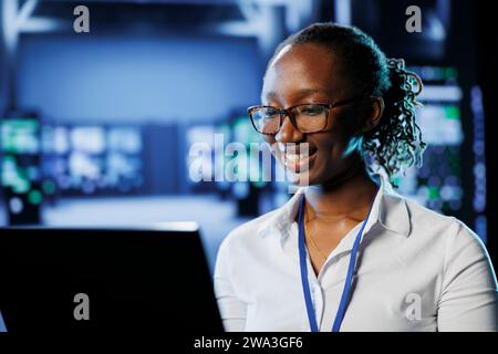 Joyful admin in high tech workplace uses laptop to prevent system overload. Competent expert between server racks ensuring enough network bandwidth for smooth operations Stock Photo
