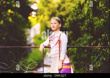 Fashion portrait of a cute little girl outdoors, wearing white sweatshirt with heart and purple bag Stock Photo