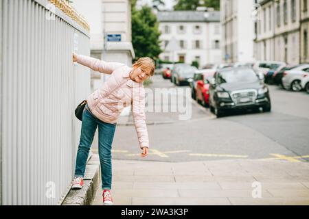 Outdoor portrait of funny little girl on the streets of Geneva, Switzerland. Child wearing soft pink padded jacket, denim jeans, and red tennis shoes Stock Photo