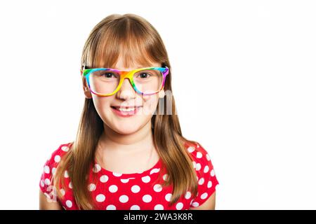 Studio portrait of adorable little 9-10 year old girl, wearing rainbow eyeglasses and red polka dot dress, standing against white background Stock Photo