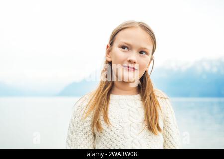 Cute little 8 year old girl playing by the lake on a very windy day, wearing warm white knitted pullover, high key image Stock Photo