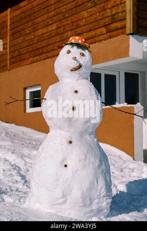 Snowman with a saucepan with red polka dots on his head stands near a wooden house Stock Photo